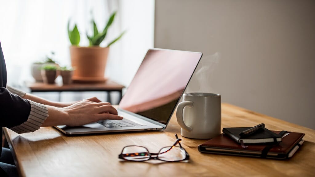 A pair of hands types on a laptop that is sitting on a table next to a pair of glasses and a cup of coffee.