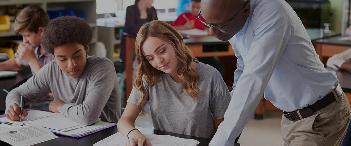 An educator leans over a desk to help a student with her schoolwork while another student sitting next to her looks on.