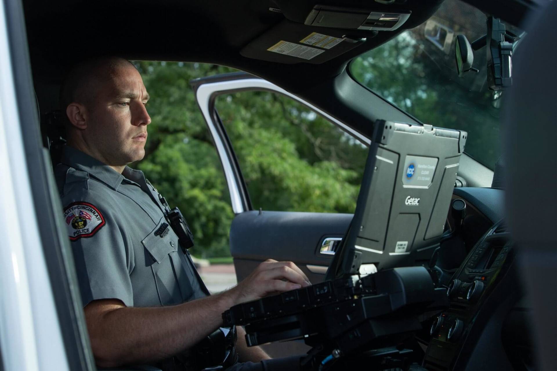 A police officer sits in his patrol car with the driver's door open as he looks at his computer and types with his right hand.