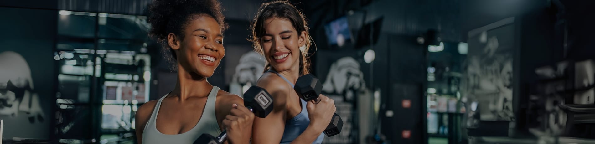Two women in athletic clothing standing back to back and smiling while lifting dumbbells.
