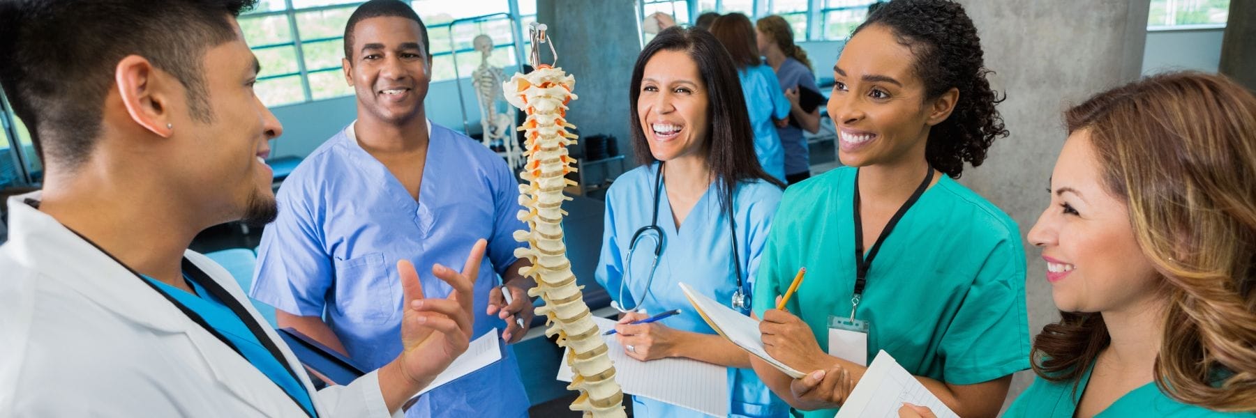 Group of nursing students and instructor discussing a spine model during a hands-on anatomy lesson in a classroom setting.
