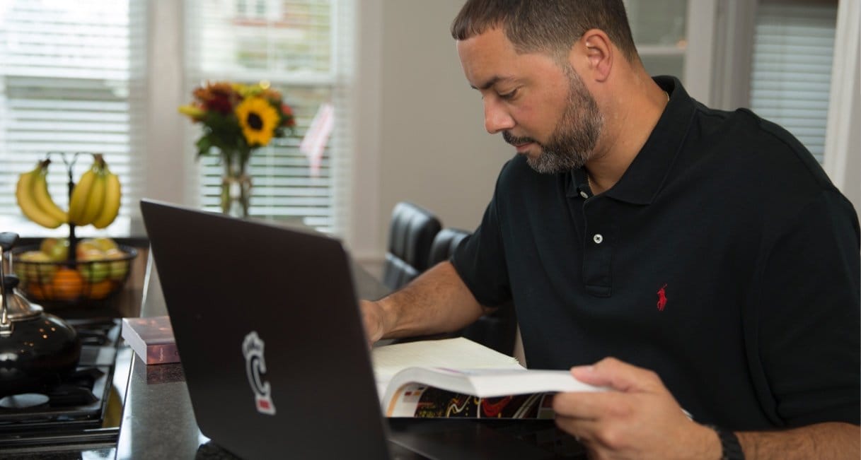 Male student reading a textbook for his online course while continuing his education