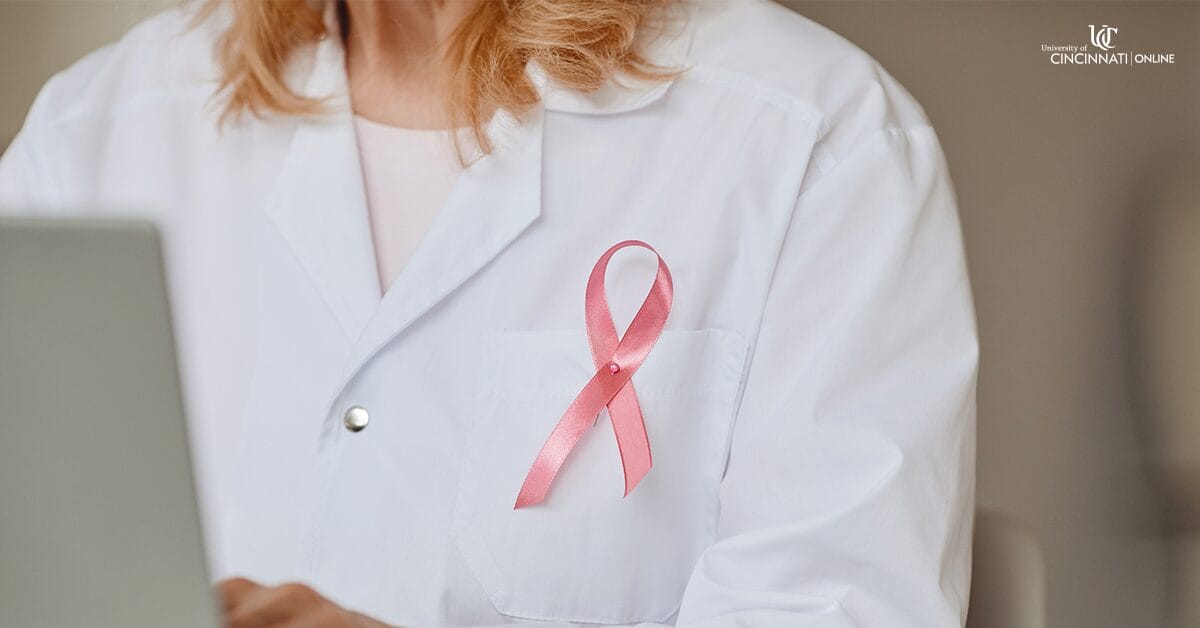 A woman sits in front of a computer and wears a white lab coat. She is visible from the neck down and has shoulder-length blonde hair. A pink cancer ribbon is pinned to her left shirt pocket.