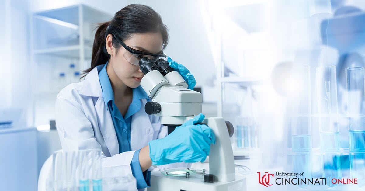 A woman with a dark ponytail wears a white lab coat and laboratory eye wear as she looks into a microscope. She is in a bright clinical laboratory setting.