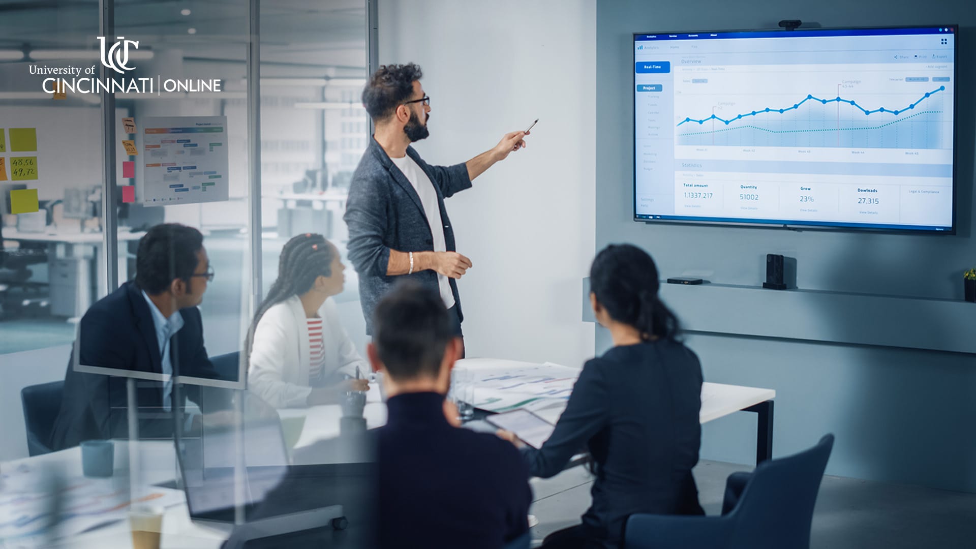 A man stands in front of a television displaying a graph. He points at it in a board room surrounded by other people who are listening to him.