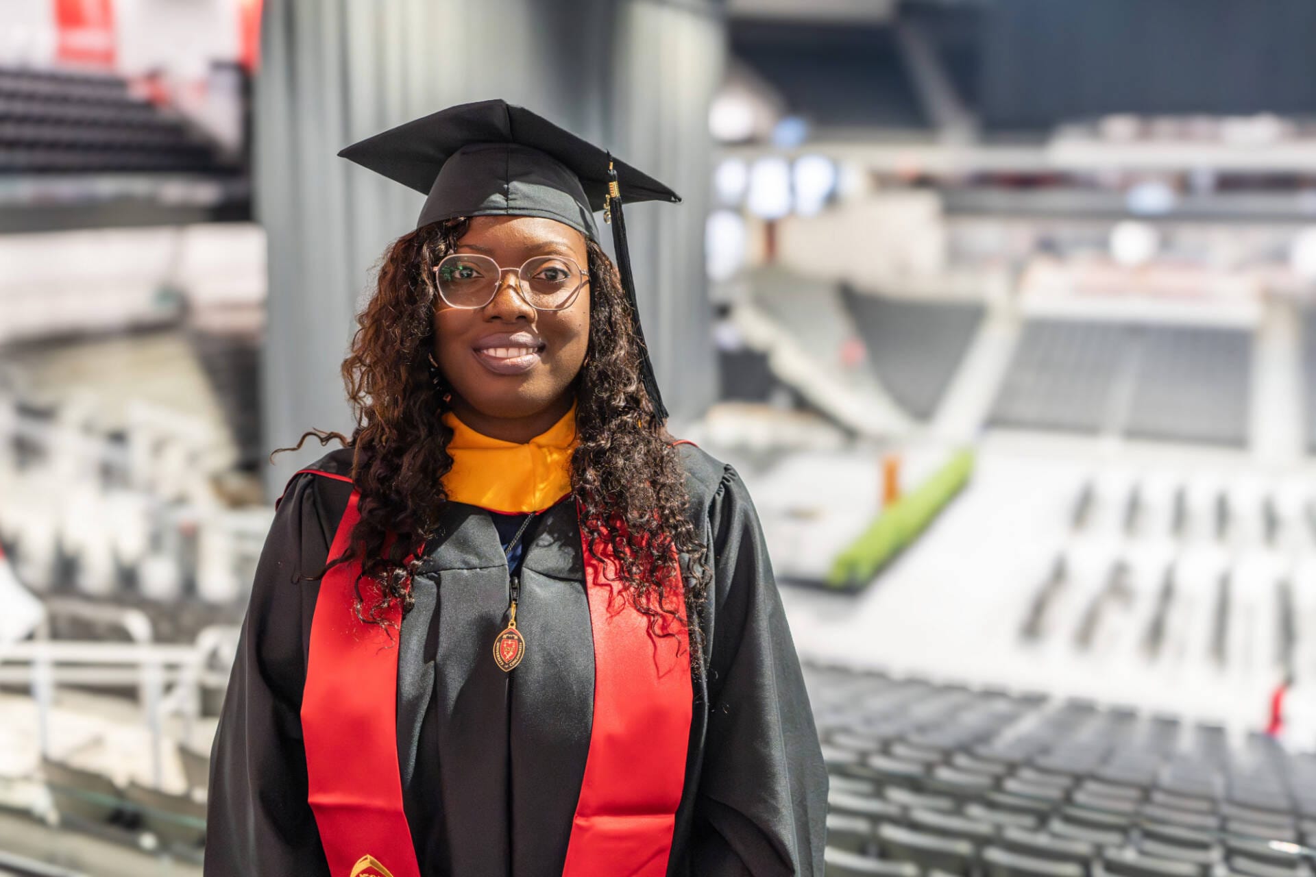 A woman stands in front of a stadium in a graduation cap, black graduation gown, and red stole. She smiles at the camera. 
