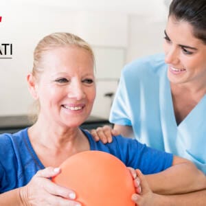 An occupational therapist rests her hand on a female patient's shoulder. The patient holds an orange exercise ball in front of her chest and smiles.