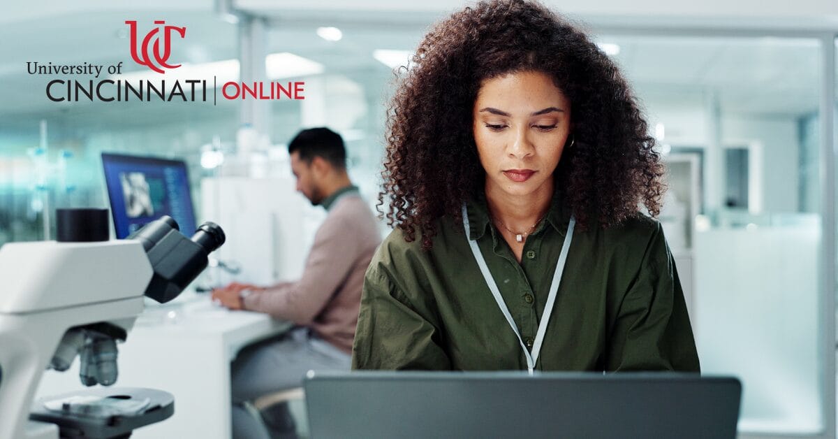 A woman sits at a laptop in a lab. She is looking down and typing. To the left is a microscope and another man typing at a computer in the background.