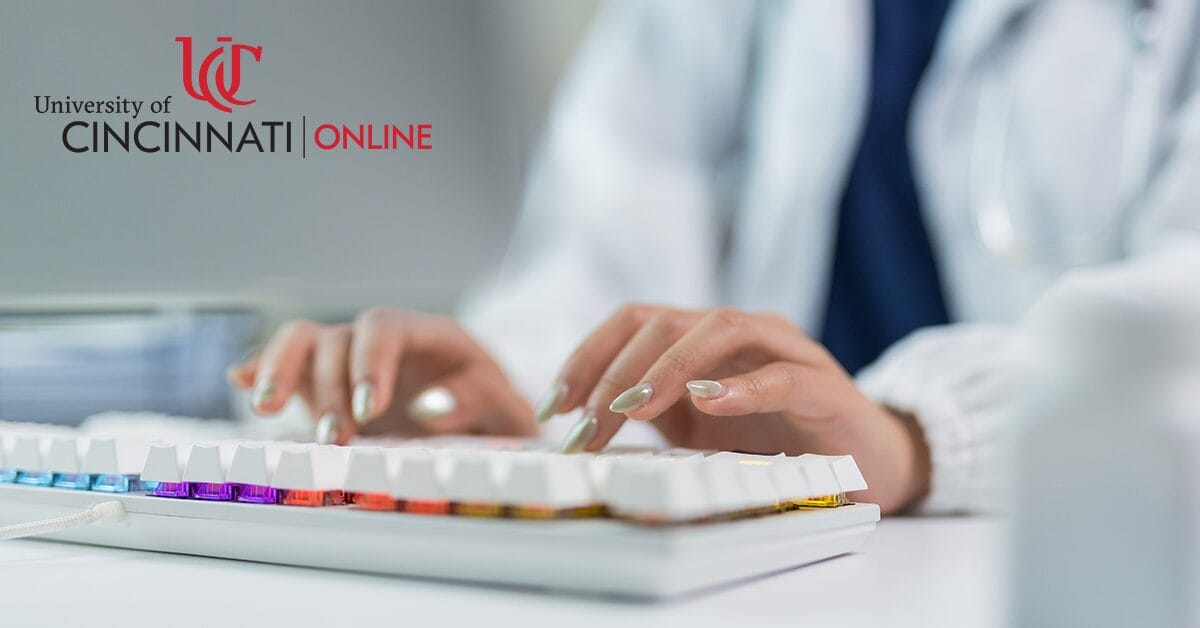 A health informatics professional types on a modern keyboard, managing health information and patient records in a clinic office. The individual wears a white lab coat and a dark shirt underneath.